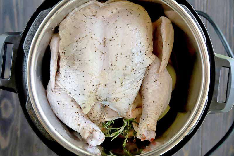 Overhead horizontal image of a raw chicken in a metal slow cooker insert, sprinkled with salt and pepper and with sprigs of fresh herbs stuffed in the cavity, on a brown wood surface.