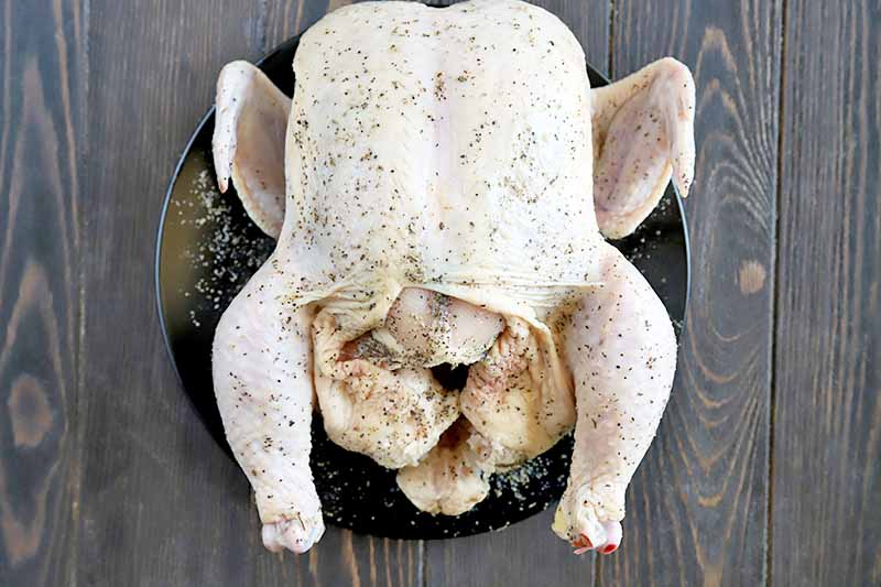Horizontal overhead image of a whole raw chicken on a small black plate, sprinkled with salt and pepper, on a brown wood background.
