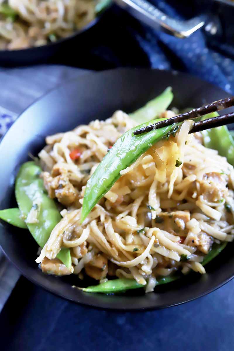 Vertical image of chopsticks holding snow peas and noodles over the entire dinner bowl of chow mein.