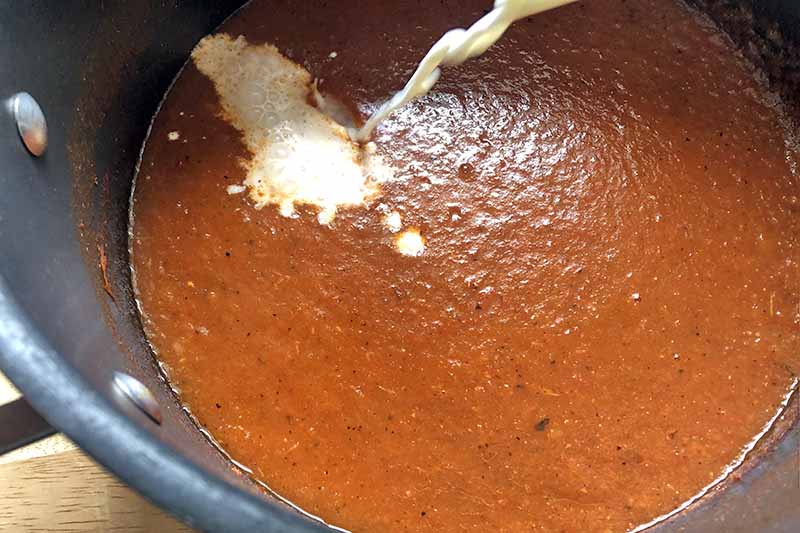 Horizontal closeup overhead image of half-and-half being poured into a red soup mixture in a saucepan, on a wood surface.