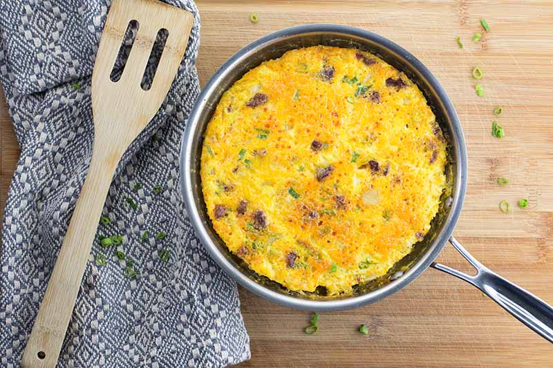 Horizontal image of a whole cooked frittata in a pan next to a spatula on a towel.