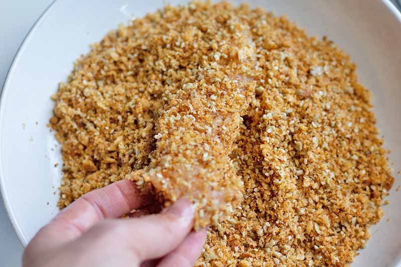 Horizontal image of coating a piece of chicken in a breadcrumb mixture.