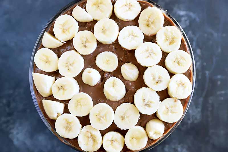 Horizontal image of banana slices evenly scattered on top of a chocolate pudding in a glass dish.