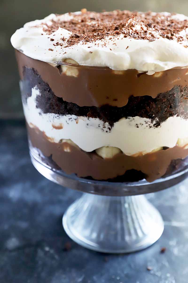 Vertical image of a large glass dish filled with alternating layers of pudding, cocoa cake, cream, and fruit chunks.