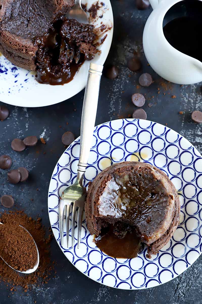Vertical top-down image of two small circular chocolate cakes with an oozing filling on blue and white plates next to candies and a spoon with cocoa powder.
