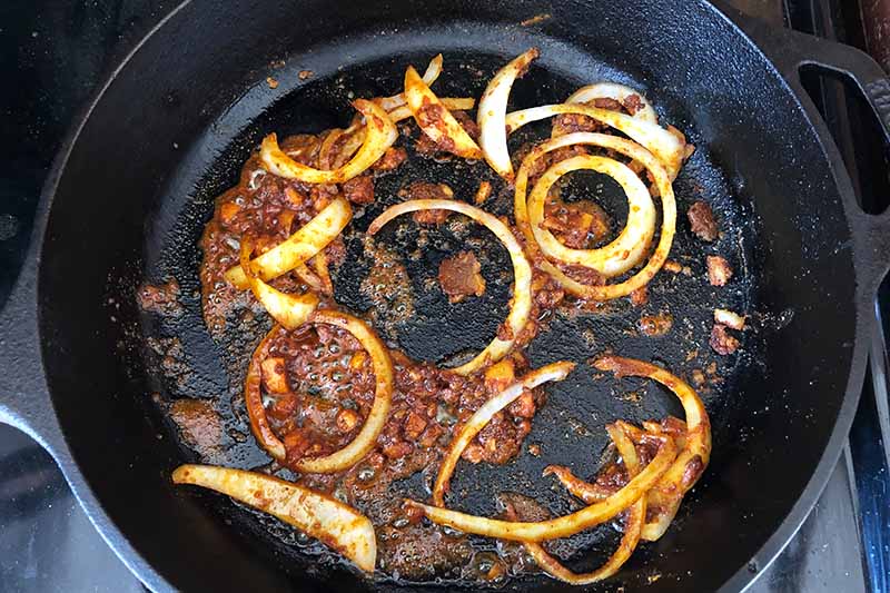Horizontal image of cooking sliced onions and spices in a pan.