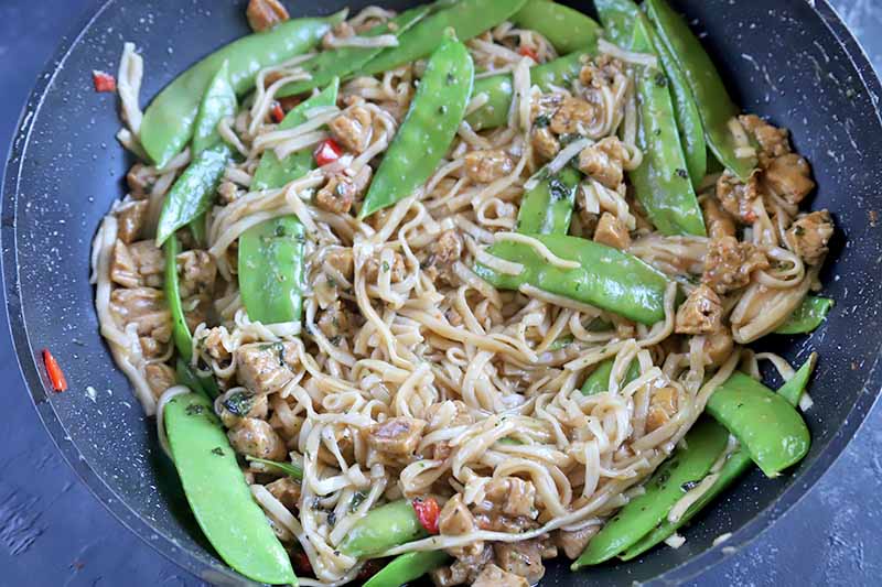 Horizontal image of noodles, snow peas, and other seasonings mixed together in a wok.