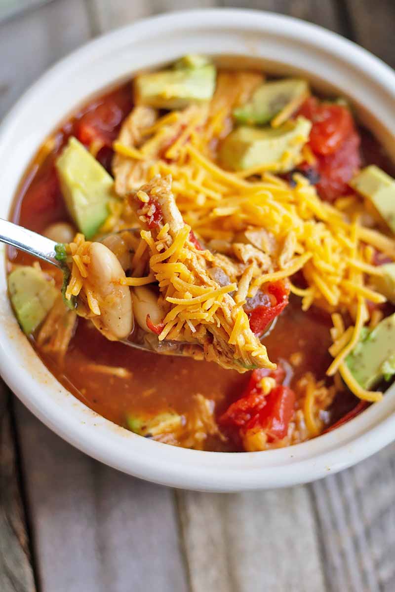 Vertical image of a white bowl filled with a tomato, bean, and turkey stew garnished with shredded cheddar cheese and chopped avocado, with a spoon holding some of it, on a wooden surface.