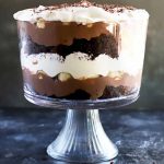 Horizontal image of a large glass serving dish with a layered dessert of pudding, whipped cream, chocolate cake, and banana slices.