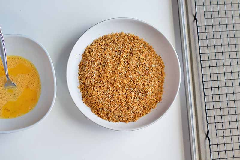 Horizontal image of a bowl of whisked eggs, a bowl of breadcrumbs, and a baking sheet.