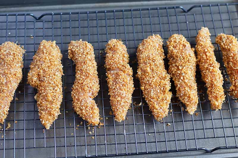Horizontal image of a row of coated unbaked tenders on a cooing rack on top of a baking sheet.