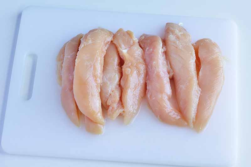 Horizontal image of a white cutting board and sliced raw poultry.