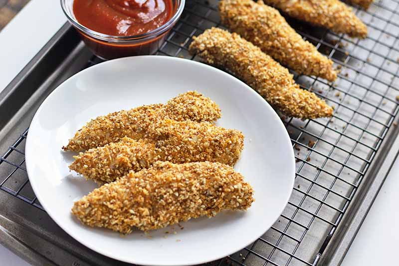 Horizontal image of three breaded chicken strips on a white plate on top of a baking sheet next to more chicken and a bowl of ketchup.