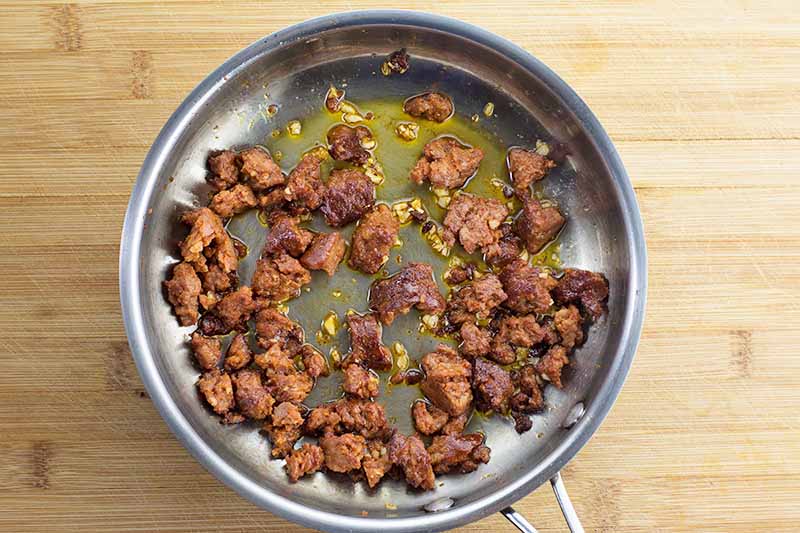 Horizontal image of cooked crumbled sausage, fat, and minced garlic in a pan.