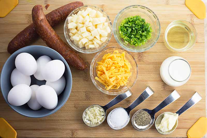 Horizontal image of seasonings in spoons and bowls, eggs in bowls, whole chorizo, shredded cheese, sliced scallions, and diced potatoes.