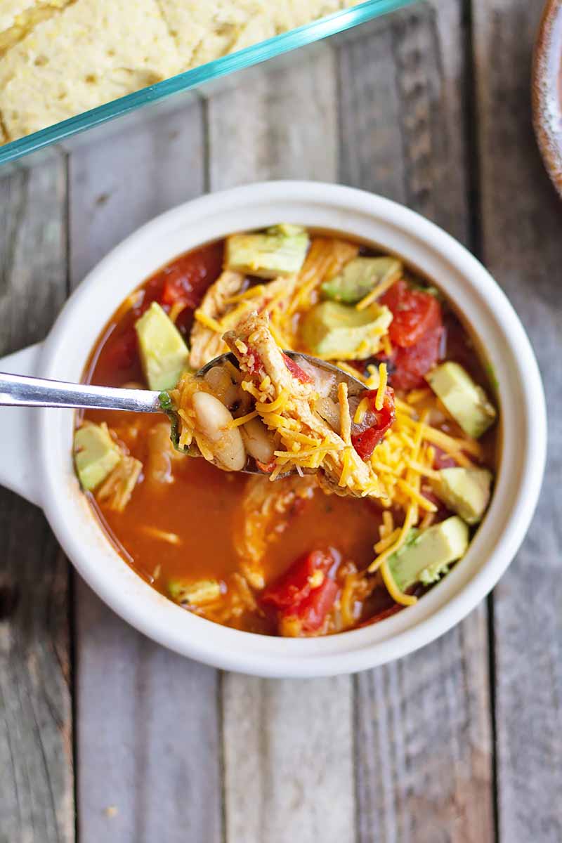 Vertical top-down image of a white handled bowl filled with a tomato and poultry stew topped with shredded cheese and avocado with a spoon dipped into it on a wooden surface next to a casserole dish with cornbread.