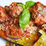 Closeup horizontal image of roasted zucchini, eggplant, and red onion with tomato sauce, meatballs, and grated cheese, garnished with a sprig of basil, on a white ceramic plate.