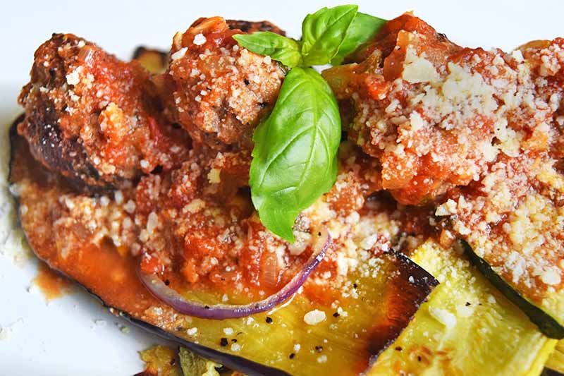 Closeup horizontal image of roasted zucchini, eggplant, and red onion with tomato sauce, meatballs, and grated cheese, garnished with a sprig of basil, on a white ceramic plate.