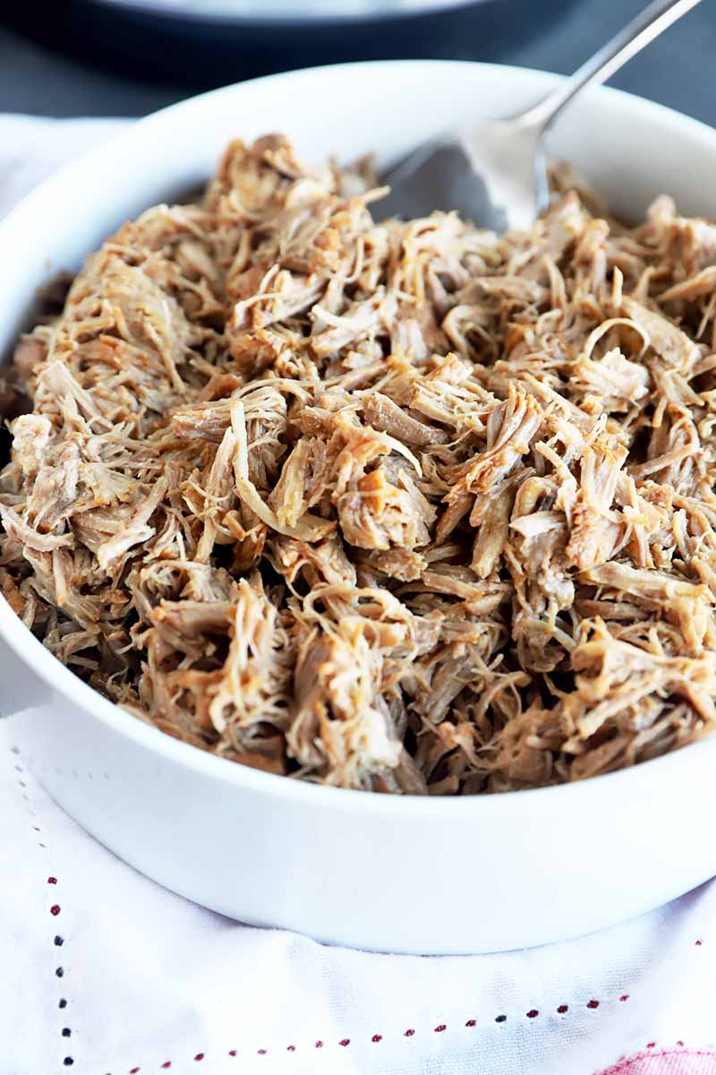 Vertical image of a large white bowl filled with a plain pulled pork and a metal fork.