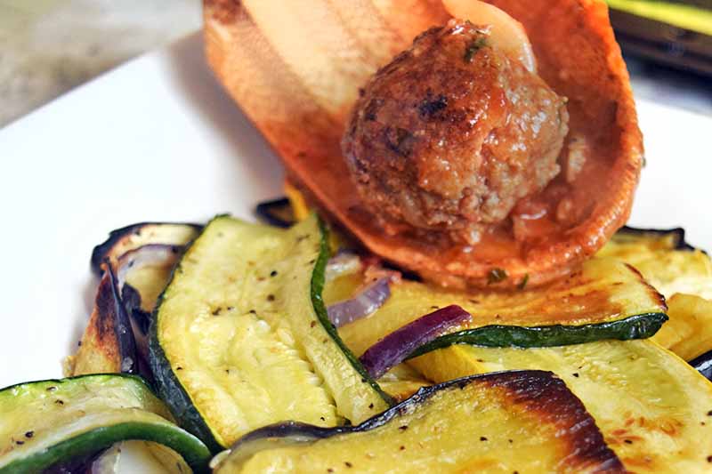 Horizontal closely cropped image of a wooden spoon transferring a meatball in red sauce onto a white plate of roasted vegetables.