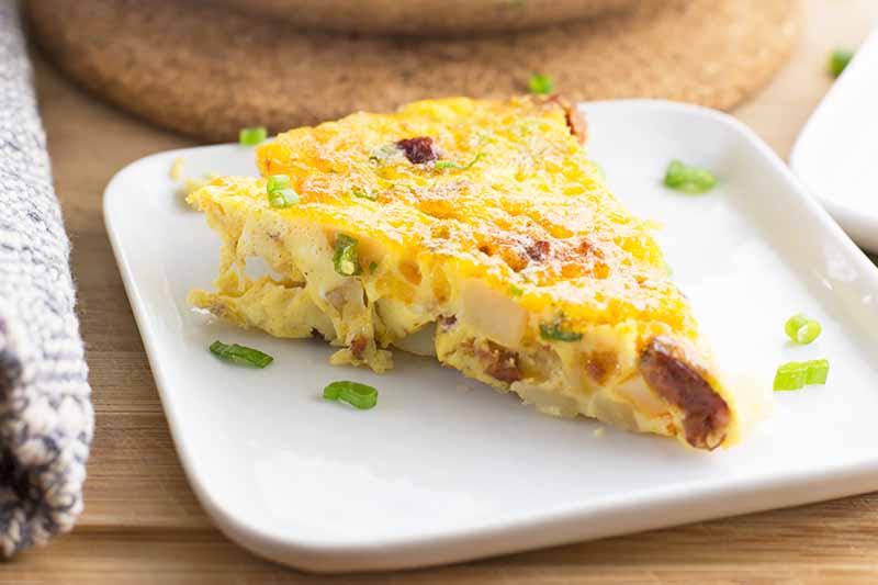 Horizontal image of a slice of an egg bake with sausage crumbles, potatoes, and scallion slices.