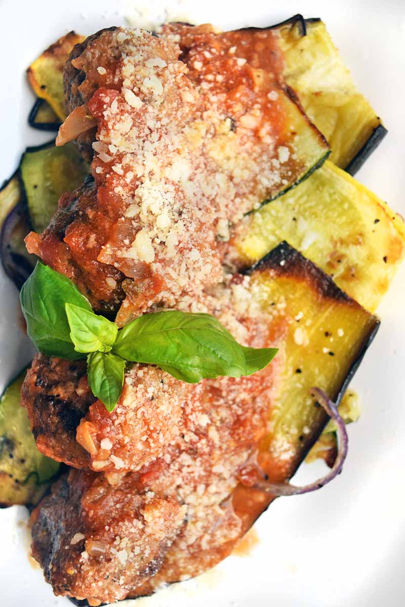 Vertical overhead image of a bed of roasted eggplant, yellow squash, and zucchini on a white plate, topped with meatballs, marinara sauce, and grated cheese, with a fresh basil sprig for garnish.
