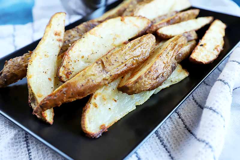 Horizontal image of a black platter with skin-on baked wedges of seasoned potatoes on top of a striped towel.