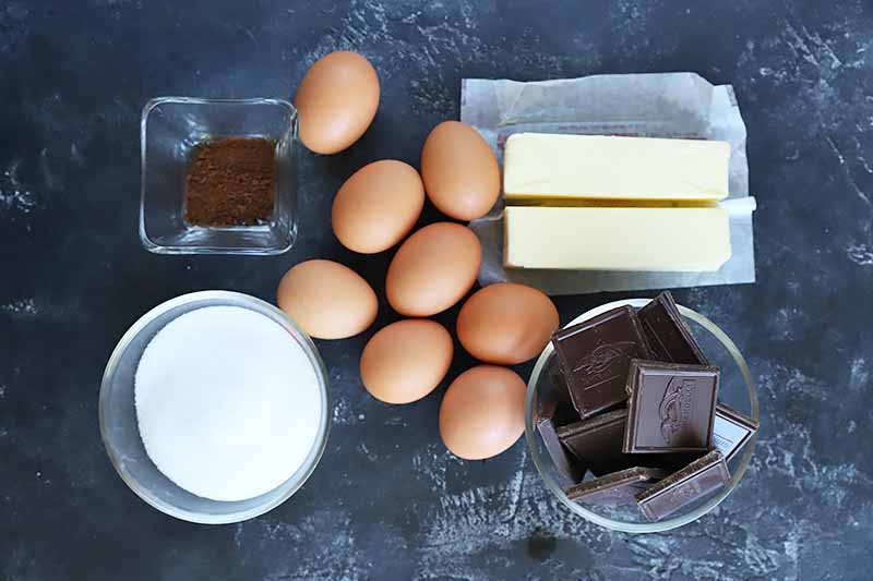 Horizontal image of eggs, chocolate, butter, and liquids in various bowls.