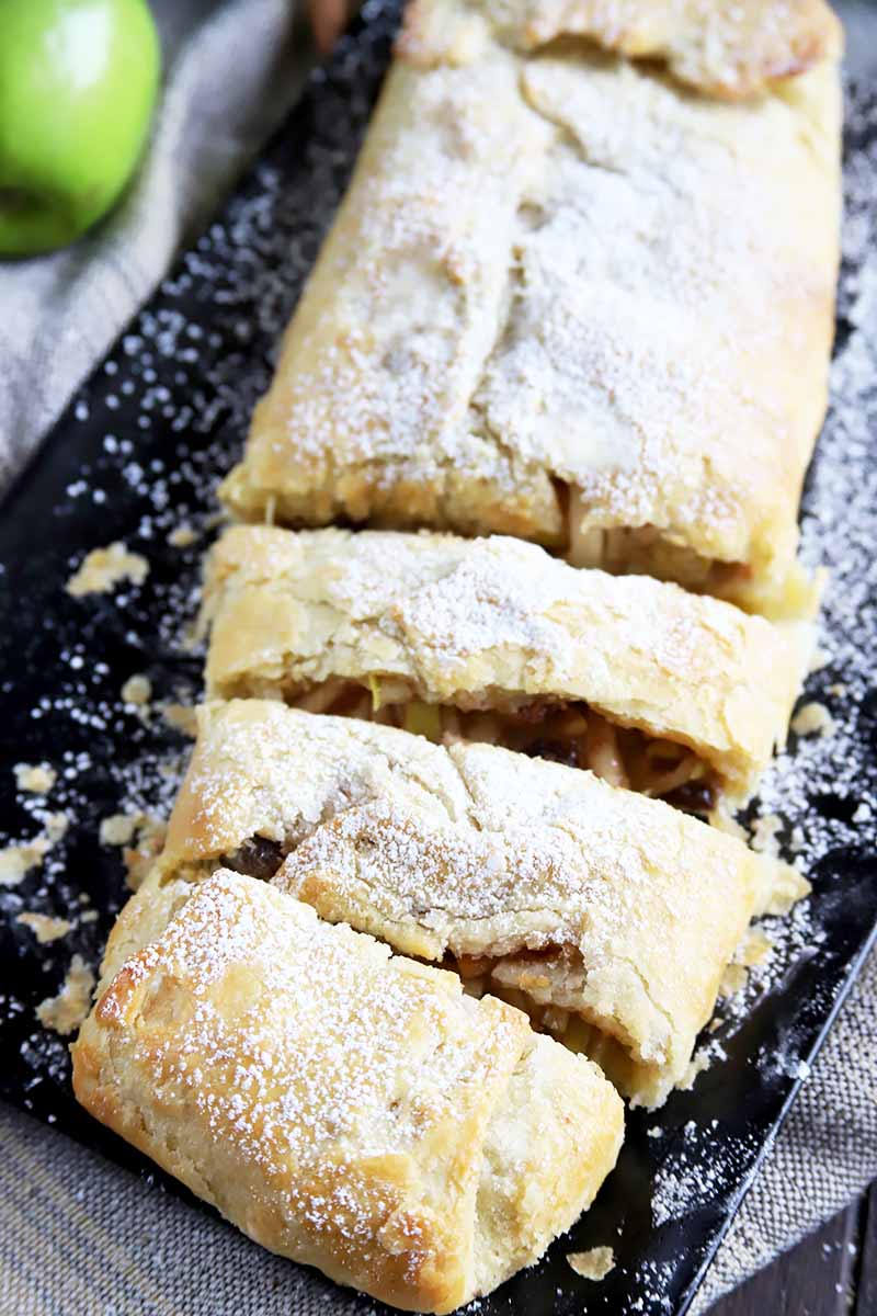 Vertical top-down image of a sliced apple strudel with a dusting of powdered sugar on a black platter on top of a towel next to a green fruit.