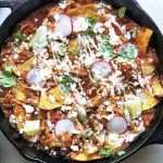 Horizontal top-down image of a black cast iron skillet filled with a chicken and sauce nacho dish topped with fresh garnishes and crema.
