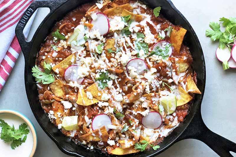 Horizontal top-down image of a black cast iron skillet filled with a chicken and sauce nacho dish topped with fresh garnishes and crema.