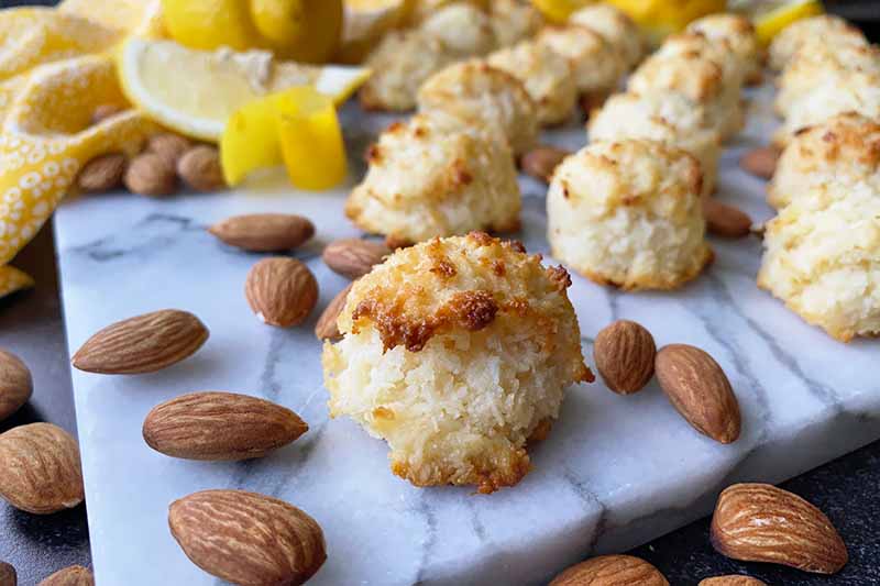 Horizontal image of one macaroon in front of others on a marble platter with whole nuts, lemon wedges, and zest in the background.