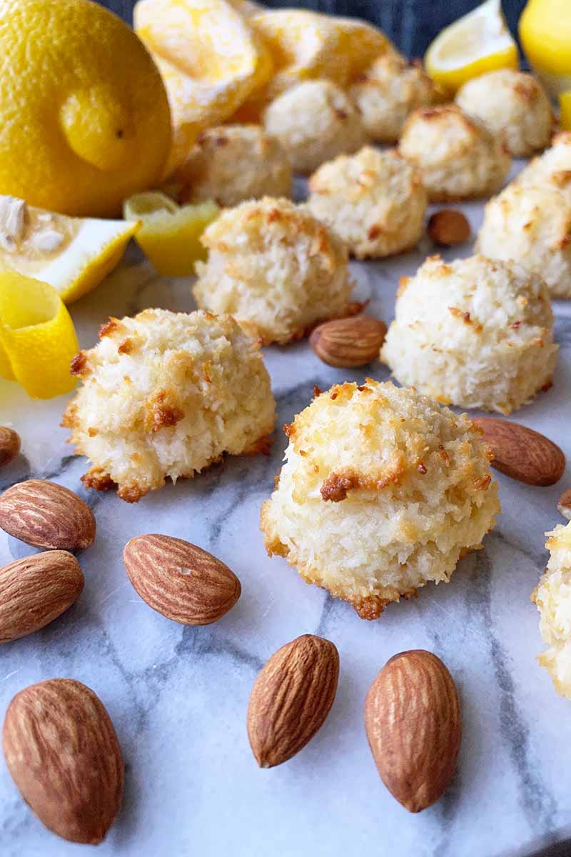 Vertical image of rows of coconut cookies on a marble board with whole nuts, citrus, citrus peels, and a yellow towel.