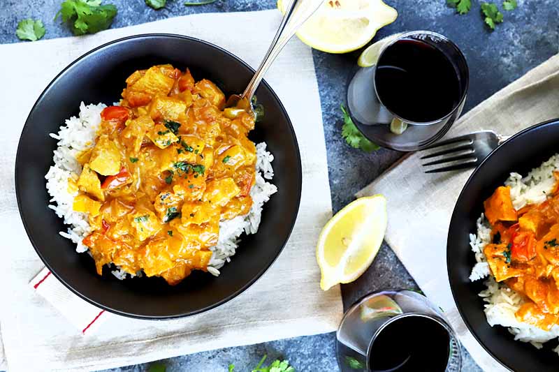 Overhead horizontal image of two black ceramic bowls of white rice topped with chicken tikka masala, with forks on off-white cloth napkins, on top of a mottled gray surface with two glasses of red wine, lemon wedges, and scattered fresh cilantro leaves.