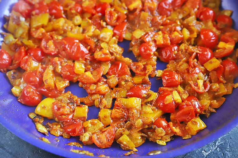 Closeup oblique overhead horizontal image of sauteed yellow bell pepper, onions, and grape tomatoes, coated in spiced, in a blue frying pan on a gray surface.