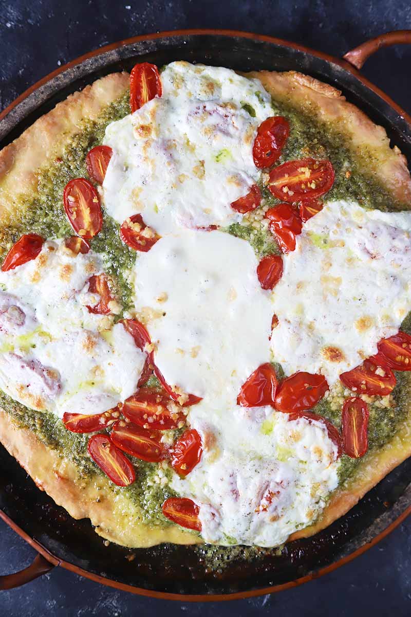 Vertical top-down image of a whole pizza with pesto, tomatoes, and melted white cheese in a large pizza stone.
