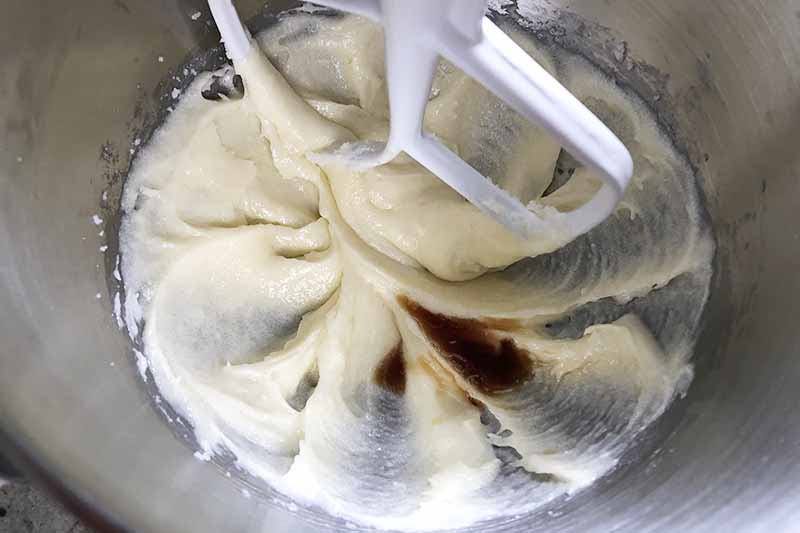 Horizontal image of mixing a thick, wet batter and vanilla extract with a white paddle attachment.