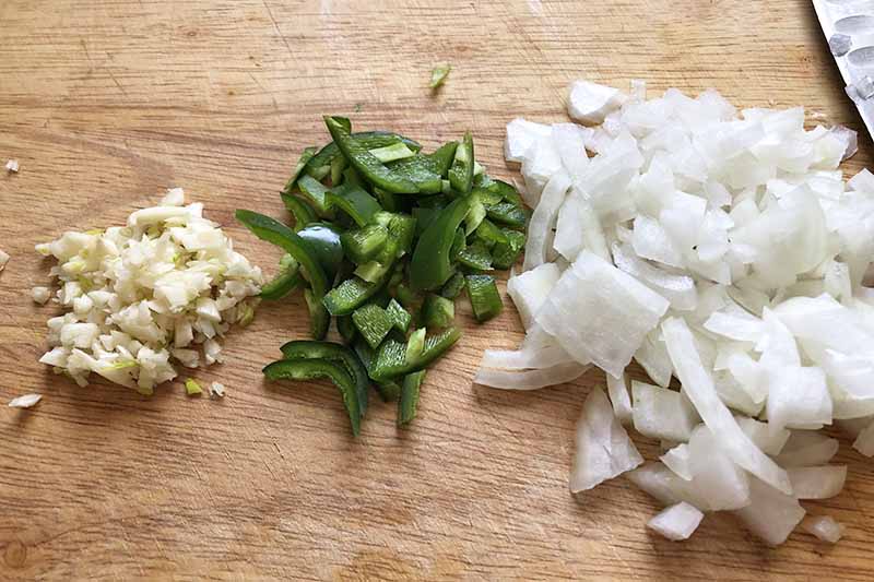 Horizontal image of chopped onions, peppers, and garlic on a wooden cutting board.