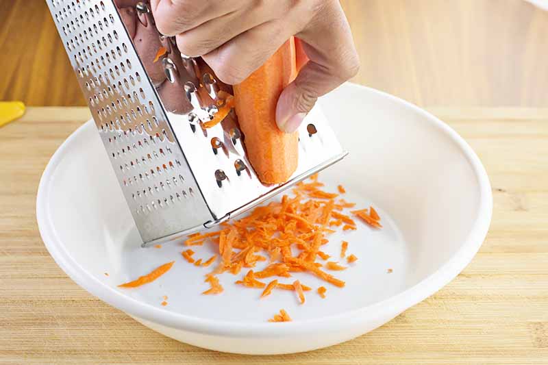 Horizontal image of grating a carrot in a white bowl with a box grater.
