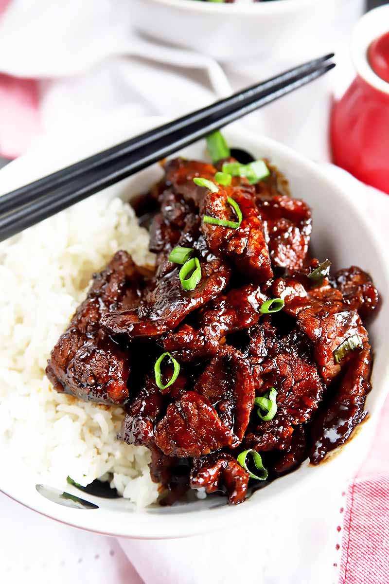 Vertical close-up image of sliced steak covered in a dark sauce with scallion garnish over a white bowl of rice next to black chopsticks.