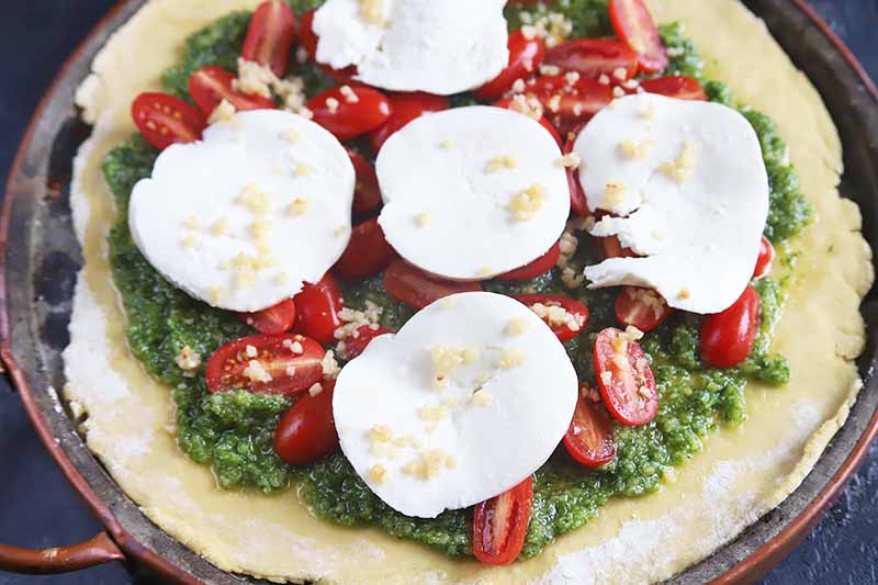 Horizontal image of an unbaked round crust with a thick green sauce, slices of tomatoes and cheese, and minced garlic.