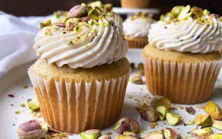 Horizontal close-up image of yellow cupcakes in white liners decorated with thick white frosting topped with pistachios.