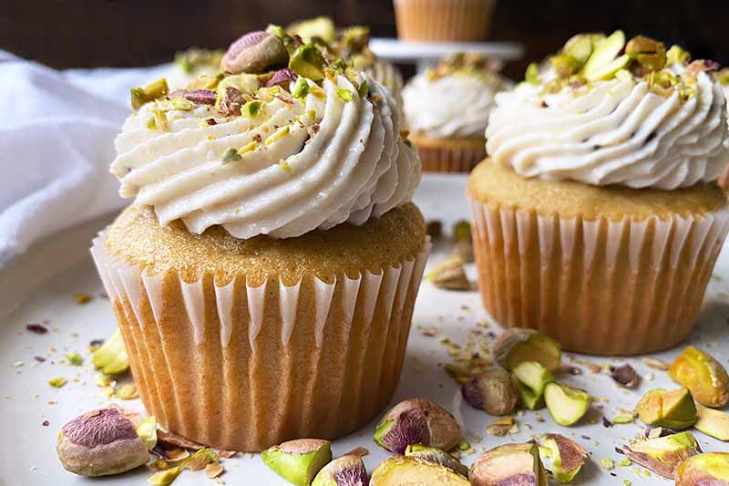Horizontal close-up image of yellow cupcakes in white liners decorated with thick white frosting topped with pistachios.