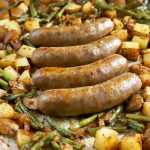 Horizontal image of four whole meat links in the middle of a tray with seasoned potatoes and beans.