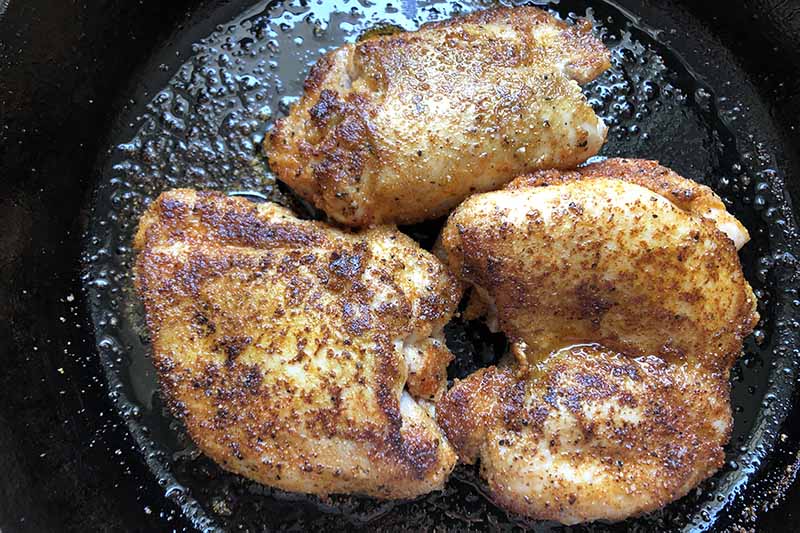 Horizontal image of seasoned pieces of poultry cooking in an oiled cast iron skillet.