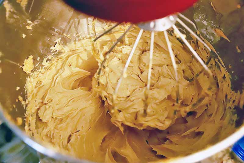 Horizontal image of a thick peanut butter frosting in a metal bowl stirred by the whisk attachment of a red stand mixer.