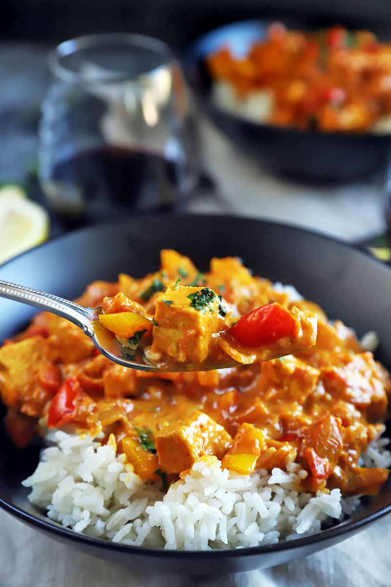 A forkful of chicken tikka masala is held above a black ceramic bowl containing more of the dish on top of a bed or rice, with another identical bowl in soft focus in the background, beside a glass of red wine and a wedge of lemon, on beige cloth napkins.