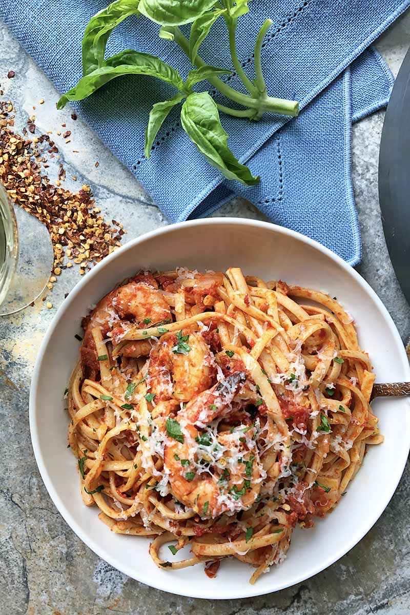 Vertical top-down image of a shrimp and linguine dish in a white bowl next to a blue apron, fresh basil leaves, a scattering of red pepper flakes, and a glass of white wine.