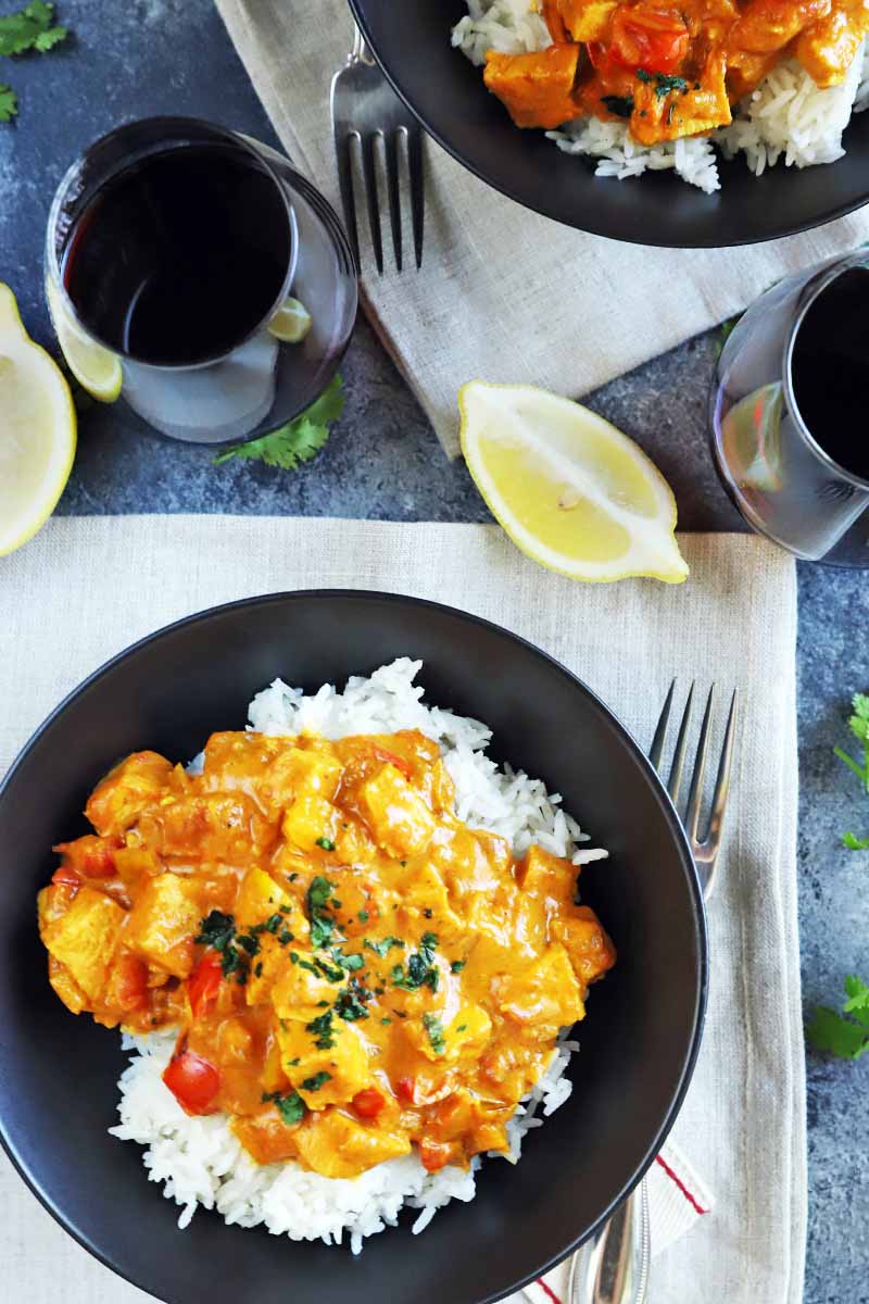 Vertical overhead image of two black ceramic bowls of chicken tikka masala atop white rice, on two off-white cloths on top of a speckled gray surface, with forks, glasses of red wine, lemon wedges, and sprigs of fresh cilantro.