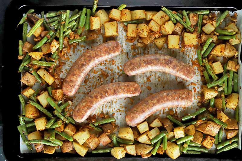 Horizontal top-down image of four uncooked meat links in the center of a sheet pan with cooked potatoes, green beans, and onions.